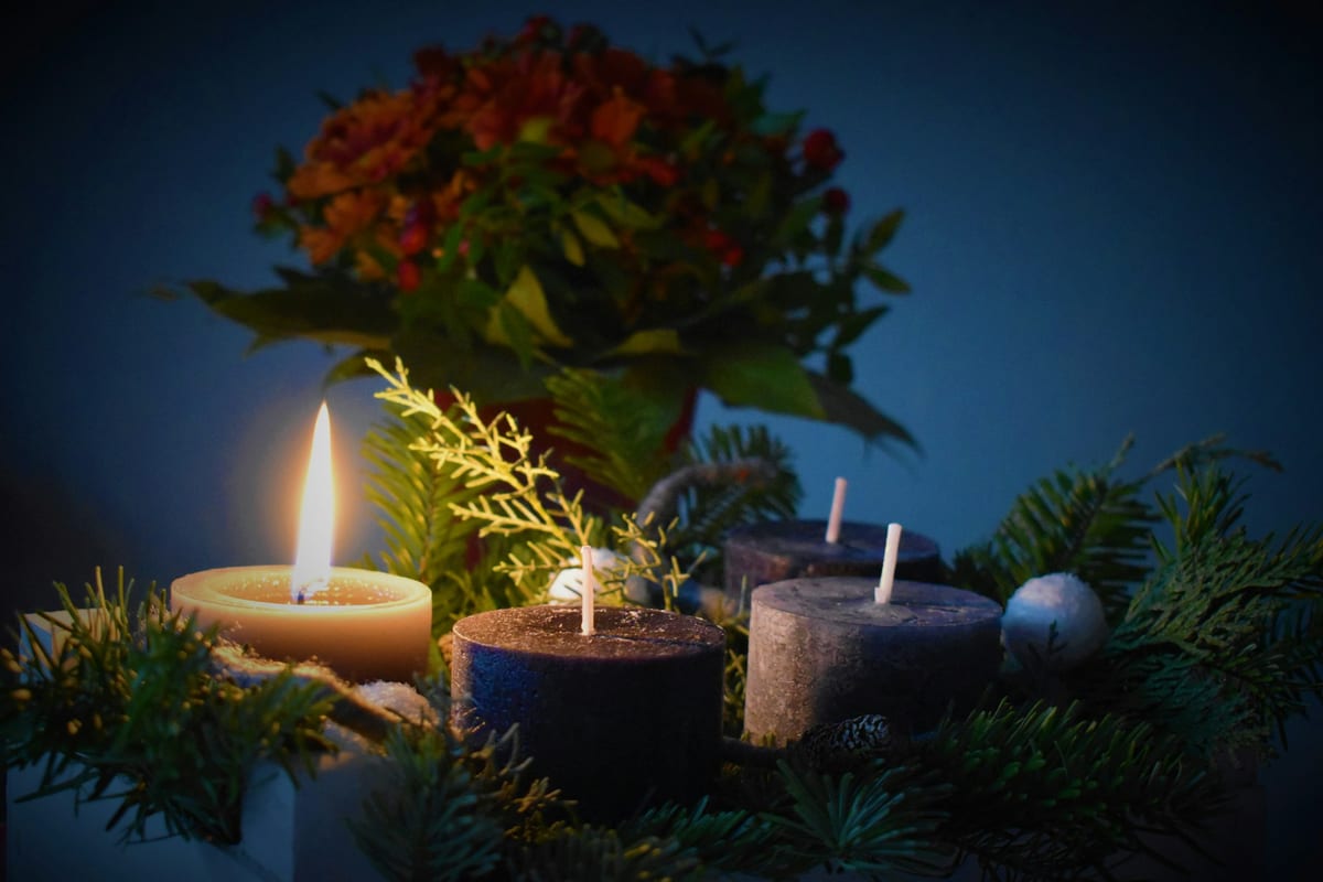 One lit candle beside three unlit candles in an evergreen wreath, in deep blue evening light