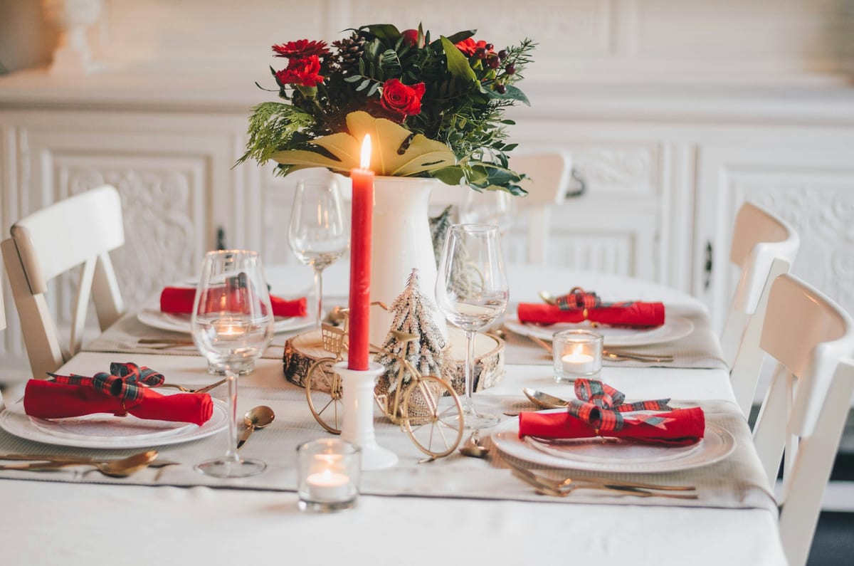 A white table set for Christmas dinner with red napkins, glasses, tea lights and a red candle beside a bouquet of roses