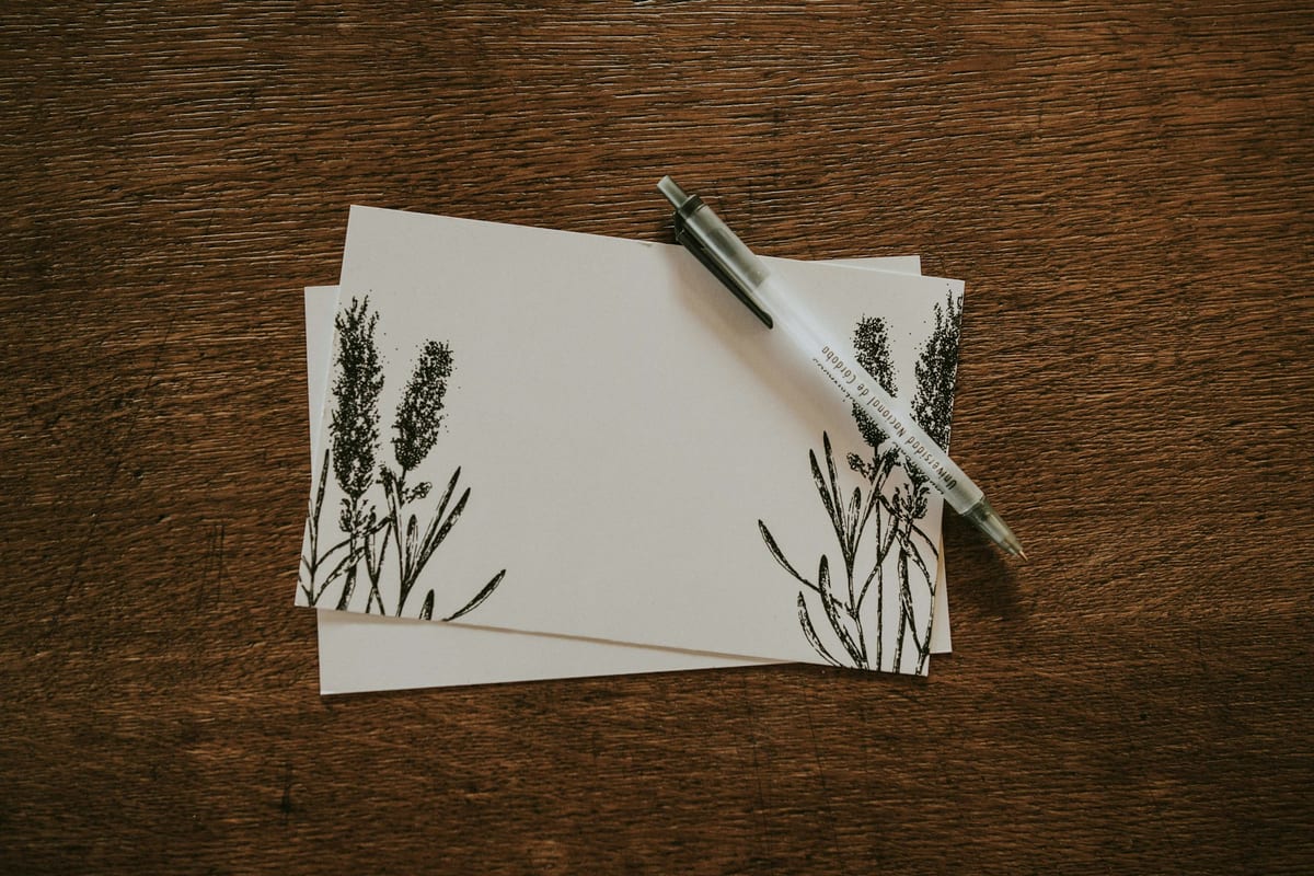 Two cream note cards with pressed-flower drawings and a white pen lying on a dark wooden table