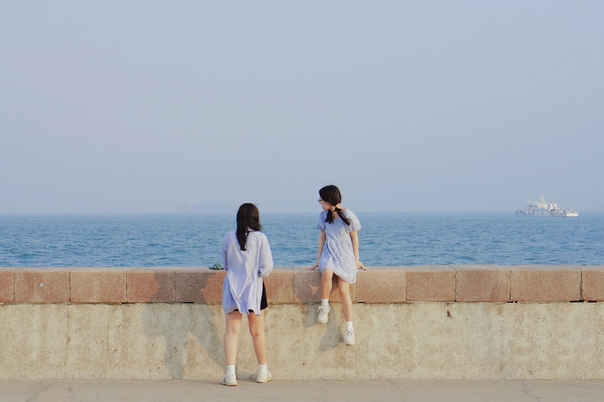Two friends at a seawall looking out over a calm sea, one standing and one sitting on the wall