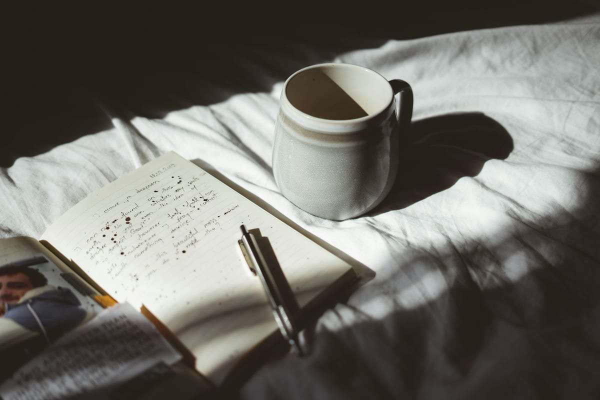 An open notebook with handwritten notes and a pen on a rumpled white bedsheet, next to a mug in morning sunlight